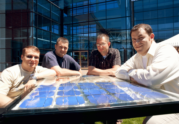 Three students post around a horizontal solar panel in front of a glass PSU building.