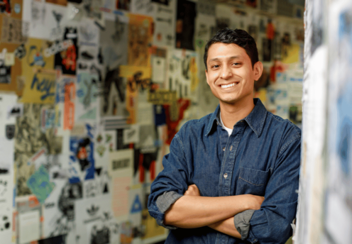 A young man wearing a casual shirt smiles in front of a wall of posters. 