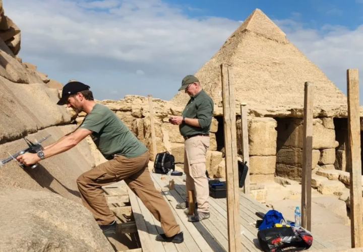 two men stand on a wooden platform attached to a Pyramid of Giza; one is scanning the pyramid with a device, the other is looking down at something in his hands