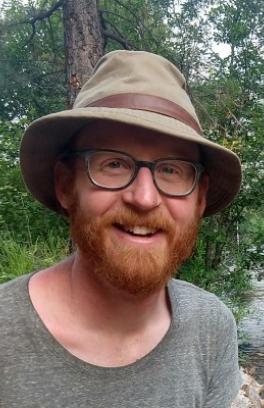 A white man with red facial hair, glasses, and a brown hat, against a backdrop rees and water