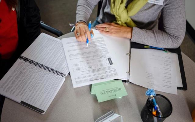 Two people sit at a table with open documents, one person pointing at a form with a pen while discussing notes.