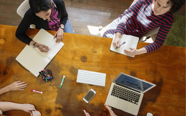 Five people sit around a table in conversation. They reference laptops and take notes.