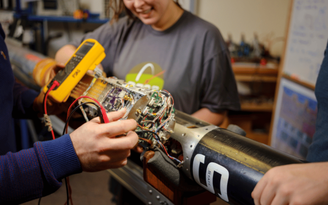 Students work on a long tubular object with dense wires. 