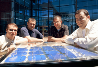 Four men stand around a solar panel in front of a glass building.