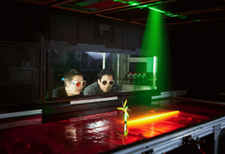 Two students wearing protective glasses watch a laser pointed at a miniature wind turbine over a tub of water.