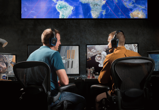 Two men hearing headphones watch screens showing maps and satellites. 