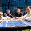 Three students post around a horizontal solar panel in front of a glass PSU building.