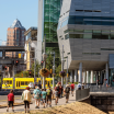 Pedestrians walk in the foreground, various buildings, public transport, and highway infrastructure cross in the background. 