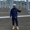 A man in work boots and a hard hat gives a thumbs up in front of an electrical substation.