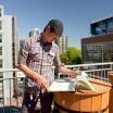 A person wearing a cap and plaid shirt reads a large open book on a rooftop with city buildings in the background.