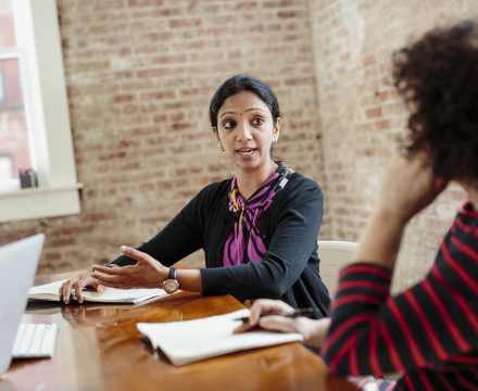 Two women talk at a conference table. They both have notepads and pens.