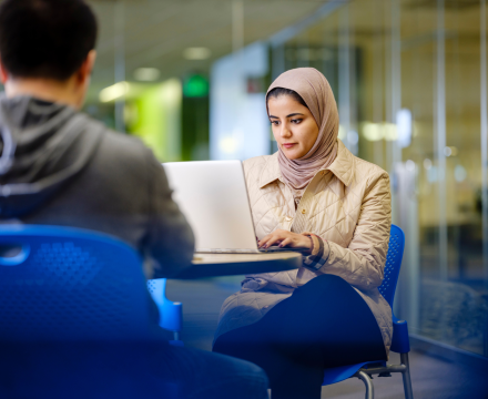 Two students work on laptops in front of a glass wall. 