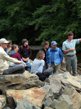 Group of people on rocks near stream. Man standing in front pointing to the distance