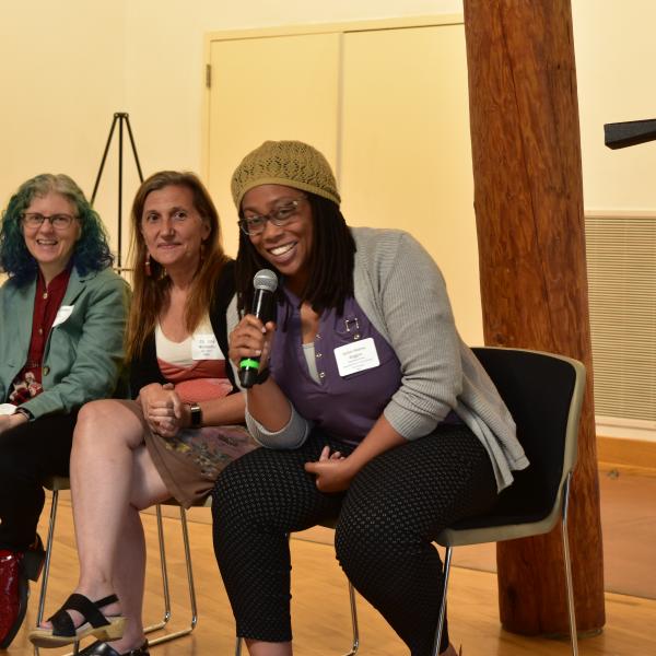 three faculty members sit in chairs on a panel, one holds the mic and is smiling at the camera