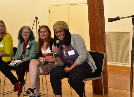 three faculty members sit in chairs on a panel, one holds the mic and is smiling at the camera