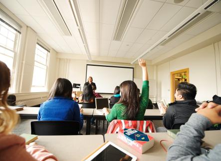 students sit in a classroom, one student raises there hand and an instructor stands at the front