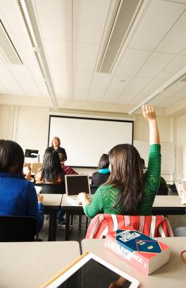 students sit in a classroom, one student raises there hand and an instructor stands at the front