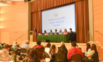 Panelists on stage with powerpoint slide behind them reading "Welcome to the 2022 Summer Research Academy."