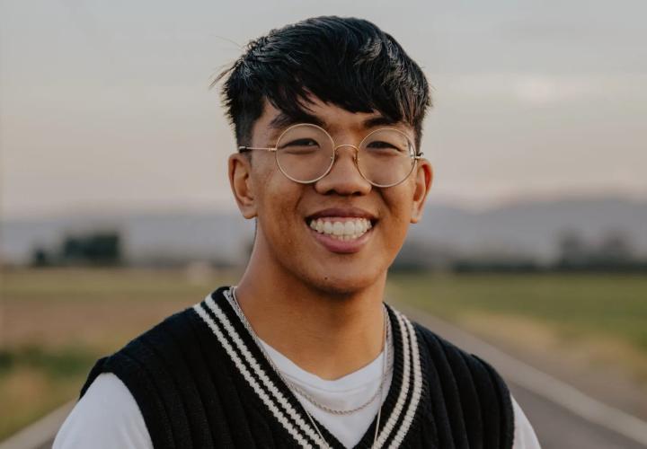 A man with glasses smiles in front of a blurred meadow and mountains