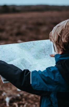 Photo of little boy from behind. He has a map open and is looking at it and open field beyond.