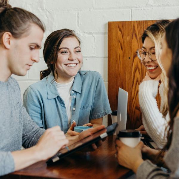 group of students sitting at a table with coffee and computers conversing and smiling at eachother