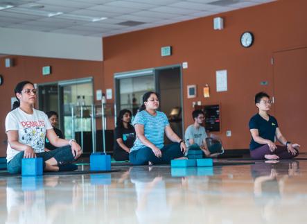 Campus Recreation - inclusive yoga and mediation in a group room. students sitting on the floor spaces out with yoga mats and stretching blocks.