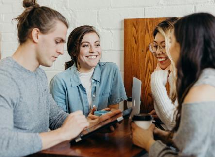 group of students sitting at a table with coffee and computers conversing and smiling at eachother