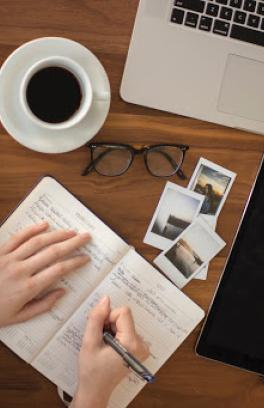 Student writing in a planner with coffee, glasses and a computer next to them.