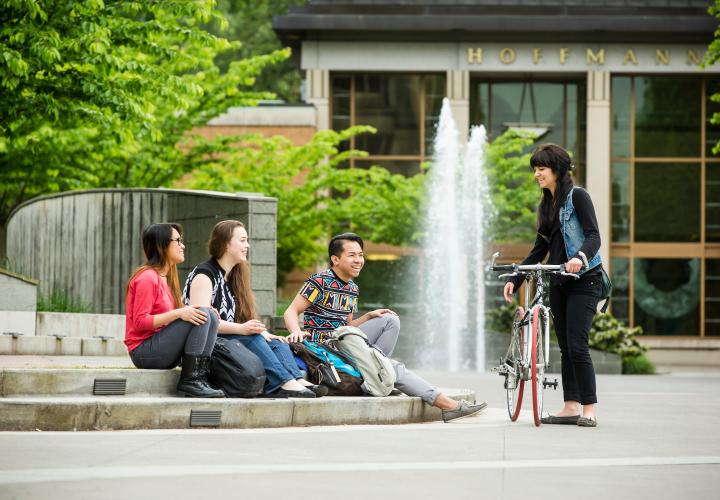 Group of four students, three sitting on steps of stage and one standing with bike