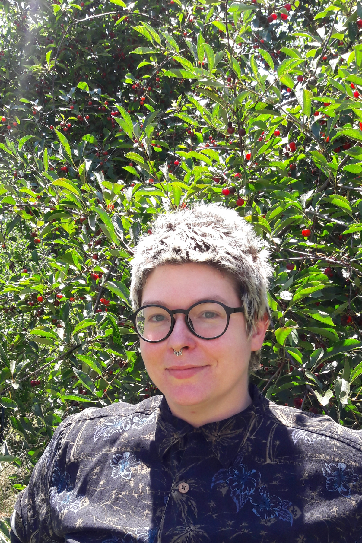 Photo of Jai Milks standing in front of green foliage with red berries. They are wearing a blue button down shirt and glasses.