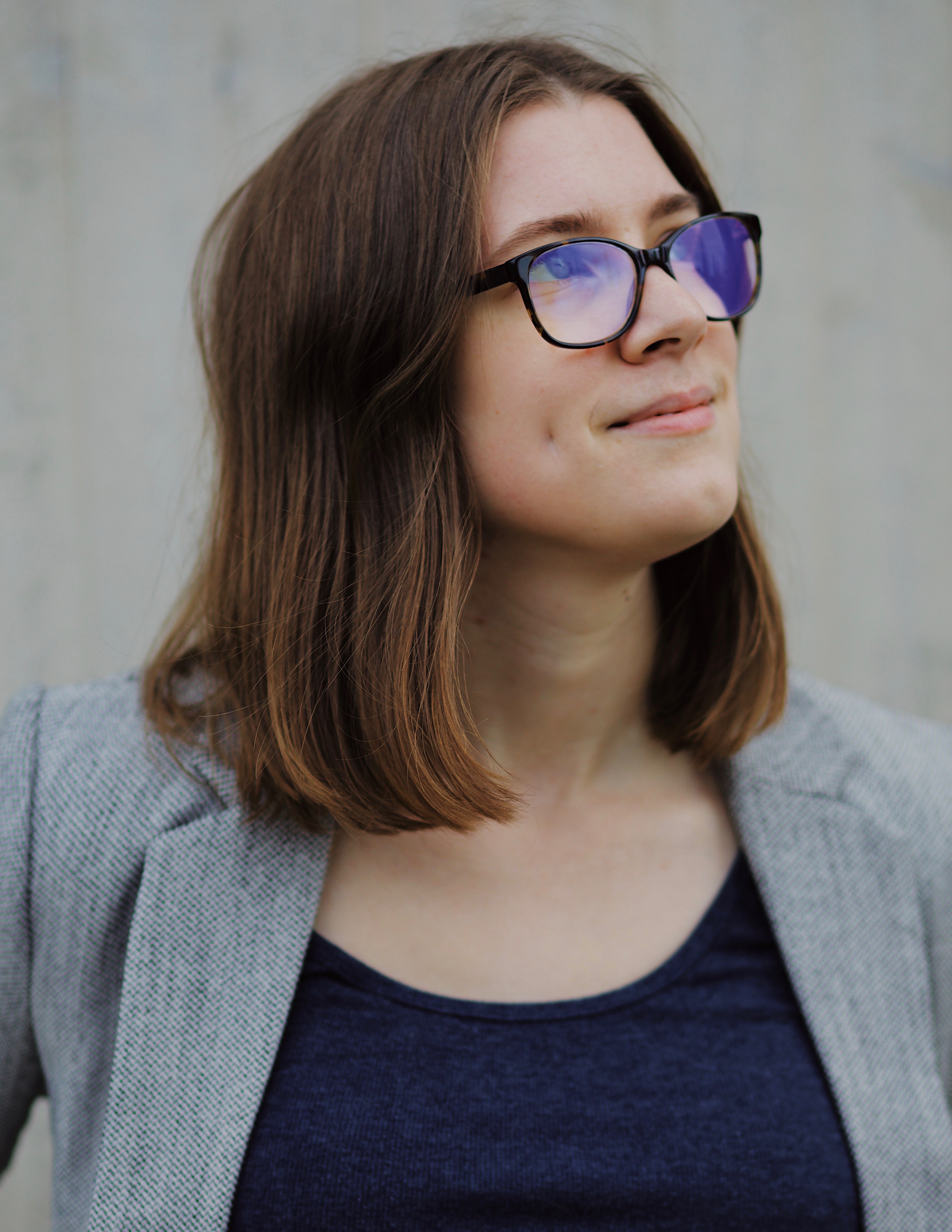 Photo of Aven Handley-Merk set against a gray background. They are wearing glasses and a gray blazer over a dark blue shirt, and they are looking up to the right. 