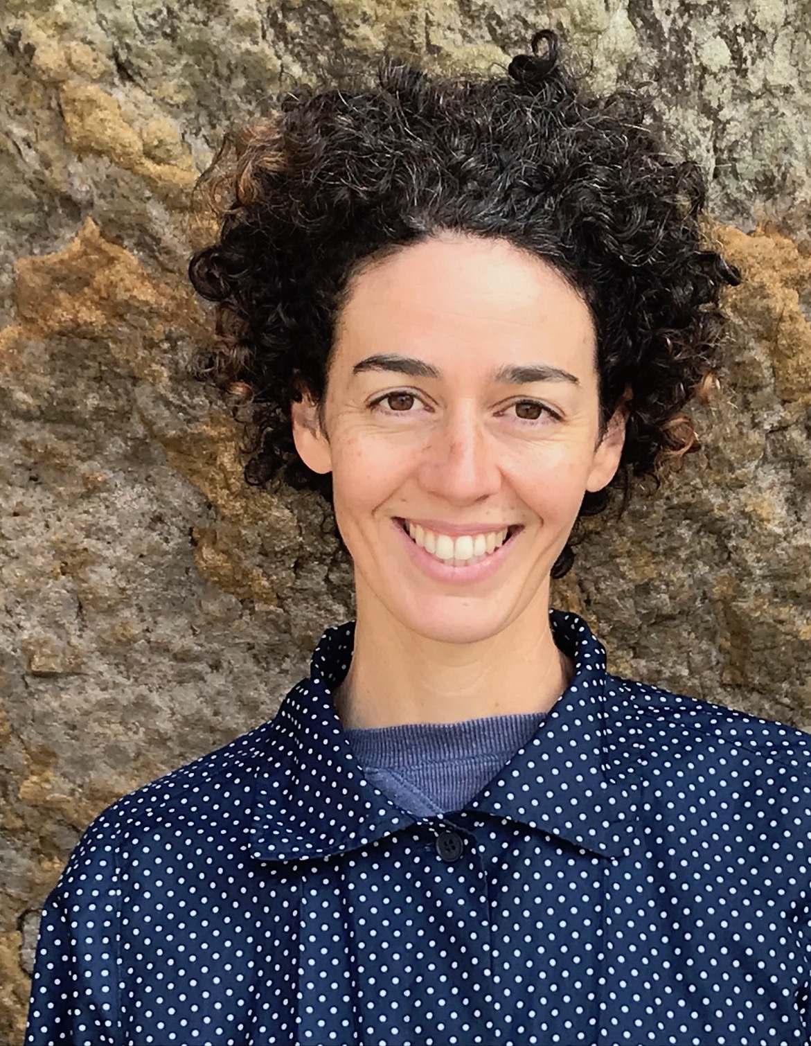 Photo of Kacy Mckinney, who is wearing a collared blue shirt with white polka dots and standing in front of a rock wall.