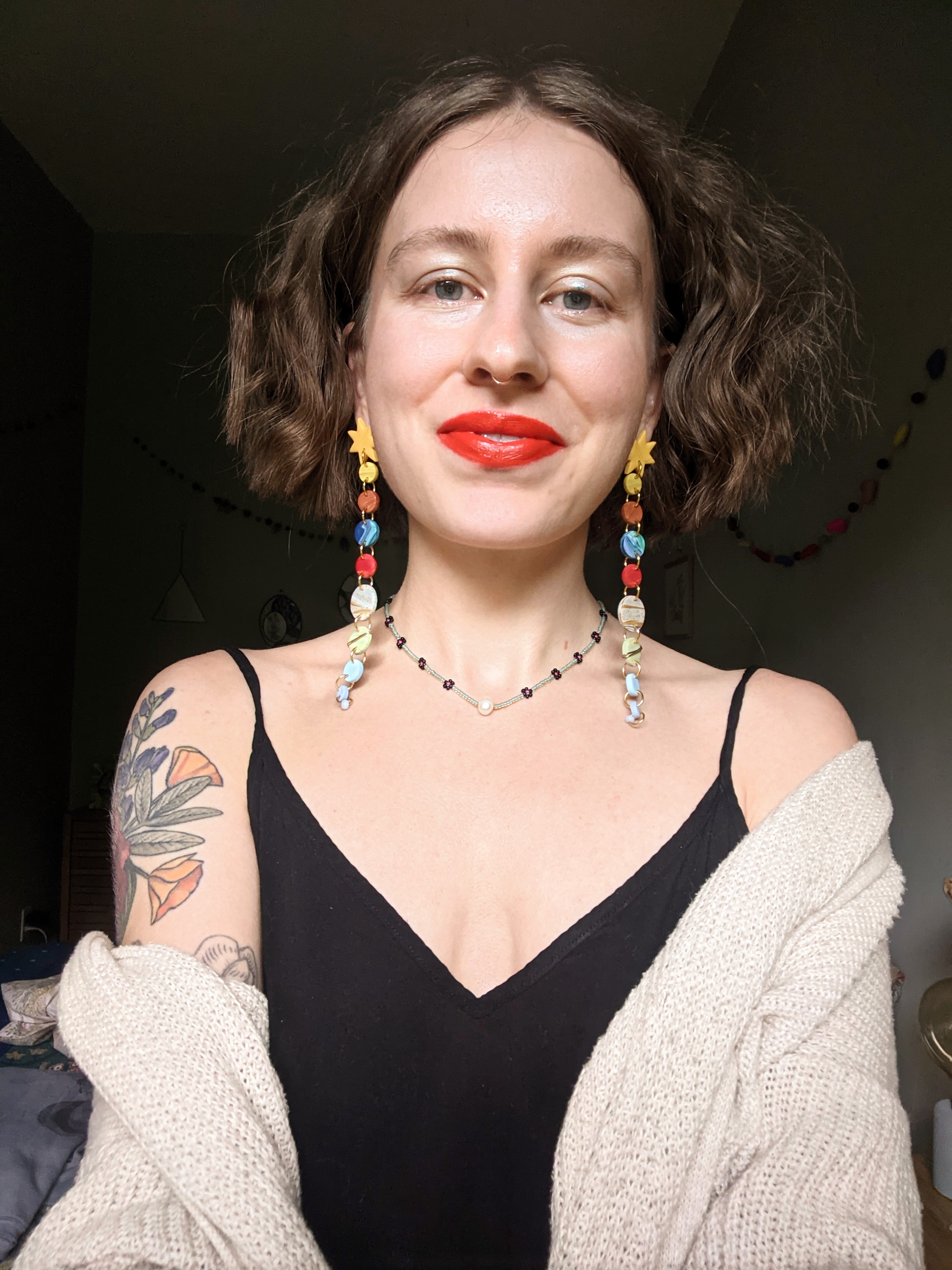 Photo of Kimberlea Ruffu set against a dark background. She is wearing a black shirt and beige sweater, and has dangly earrings and bright red lipstick. 