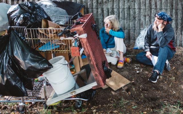 Two interviewers sit on the ground, speaking with the hidden interviewee who is behind a shopping cart of belongings.