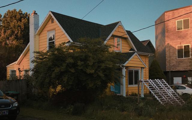 Photo of a bright yellow, two story house with a black roof against a blue sky. A short, wide tree covers much of the view of the house.