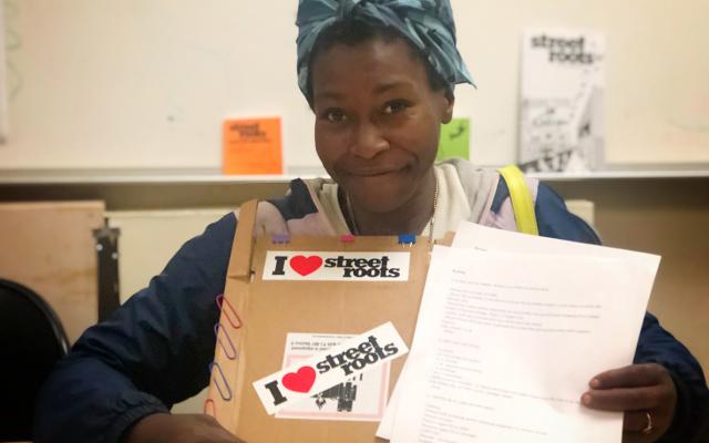 Photo of Mari Louden. She is smiling and wearing a blue scarf on her head and holding a homemade clipboard with stickers that say "I love Street Roots".