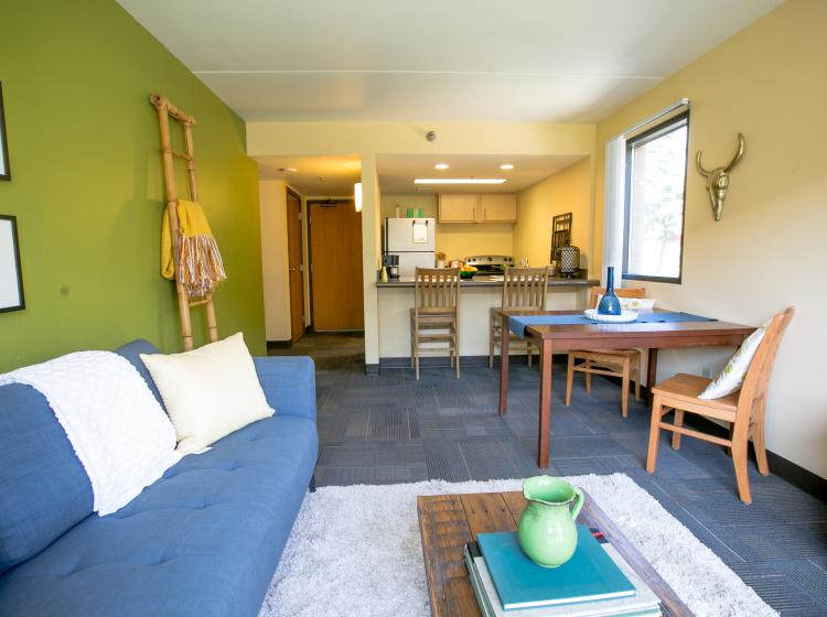 Interior picture of Blumel living space and kitchen. A couch and coffee table are in the foreground, kitchen table, barstools, and kitchen are towards the back.