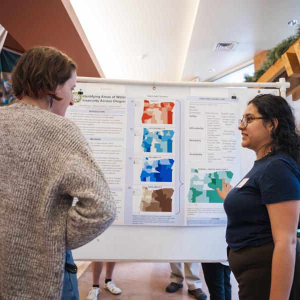 a female presenting student talking about their project with another person. They are standing in front of a research poster. 