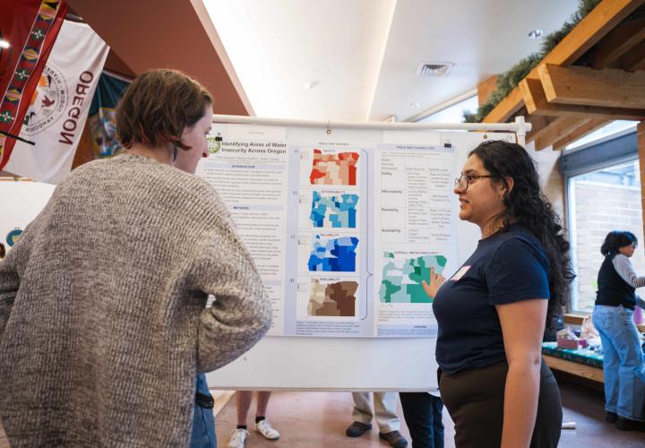 a female presenting student talking about their project with another person. They are standing in front of a research poster. 