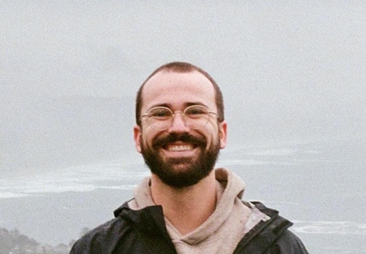 White male with glasses and a dark jacket stands in front of a foggy seashore vista.