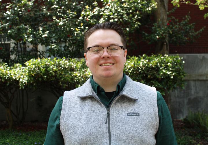 White male with glasses and a gray fleece vest smiles in front of a green hedge.