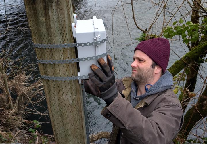 A man in a warm hat, brown jacket and gloves attaches a white box to a large post next to a river.