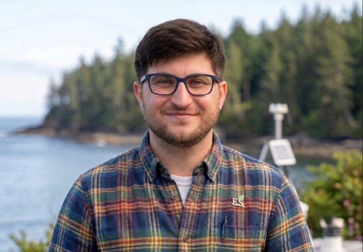 A man with brown hair and glasses in a colorful plaid shirt with trees and water in the background.