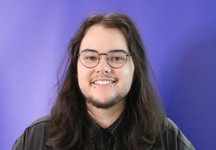 A man with long brown hair and glasses in a black shirt against a purple background.