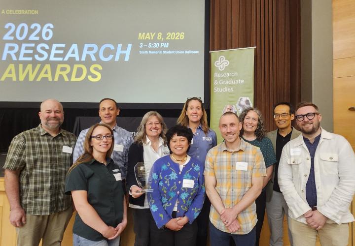 A group of ten people pose together holding a glass trophy, standing in front of a screen reading "2026 Research Awards: A Celebration, May 8, 2026," at PSU's Research Awards Ceremony.