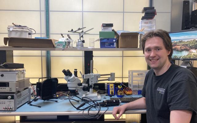 A white man sits at a lab bench surrounded by a microscope and electronic equipment