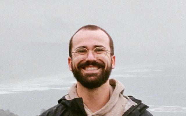 White male with glasses and a dark jacket stands in front of a foggy seashore vista.