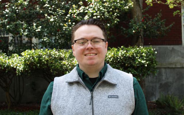 White male with glasses and a gray fleece vest smiles in front of a green hedge.