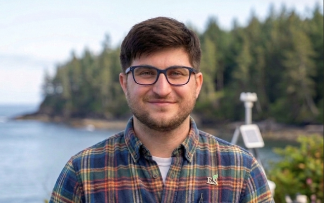 A man with brown hair and glasses in a colorful plaid shirt with trees and water in the background.