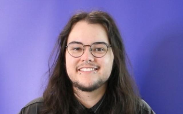 A man with long brown hair and glasses in a black shirt against a purple background.